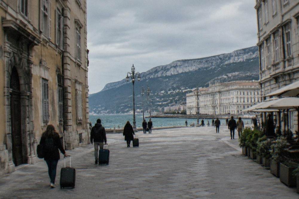 Passengers with luggage walking past the grand seafront square of Trieste on a bright day