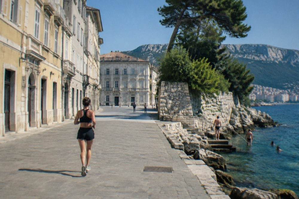 A rocky coastline with pine trees above clear water and people entering the sea from stone steps