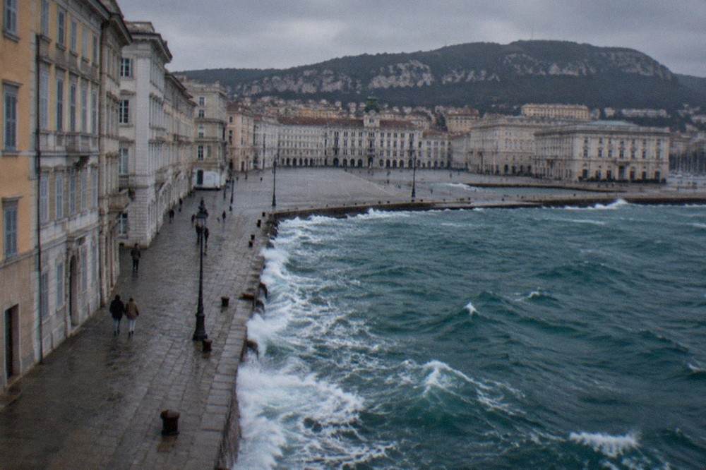 Waves breaking along the Trieste waterfront seafront on a windy grey day, with the square and historic buildings behind