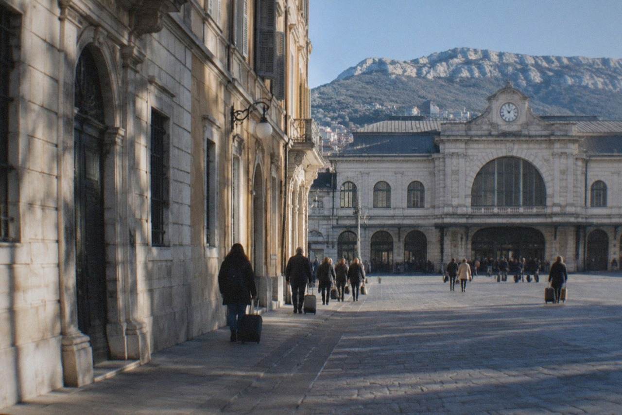 The front of Trieste Centrale railway station with its stone facade and passengers with luggage outside