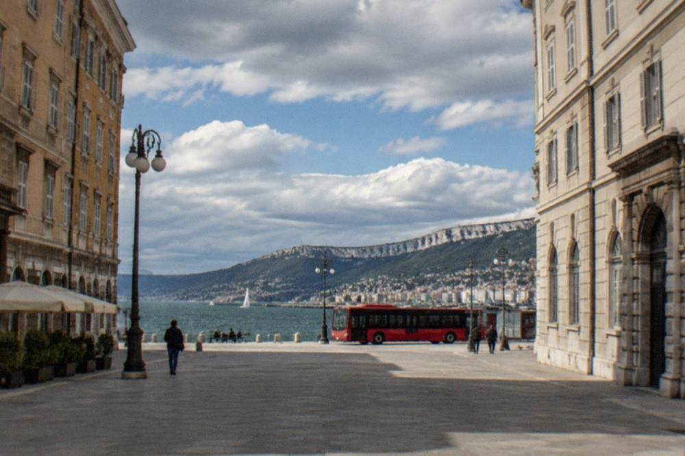 A red Trieste city bus parked at a stop with the seafront and hills behind it