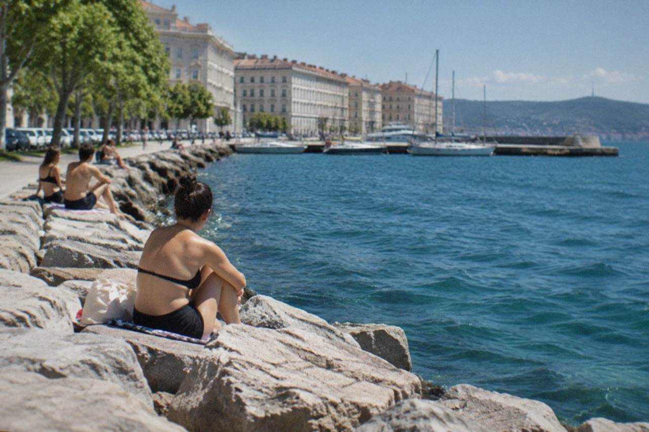 People relaxing on a rocky seafront promenade along the Gulf of Trieste on a sunny day