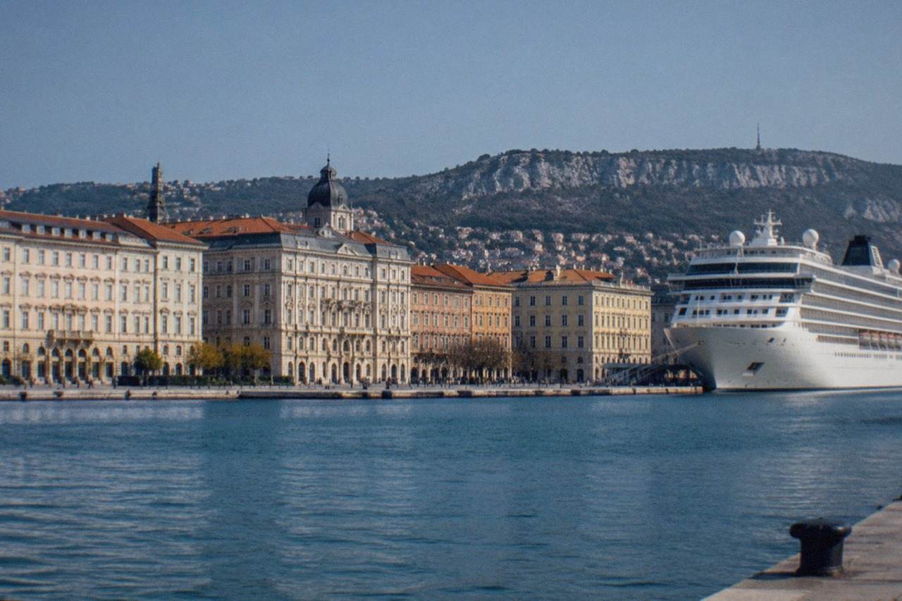 A cruise ship berthed beside the ornate waterfront buildings of Trieste, Italy, seen across the harbour on a clear day.
