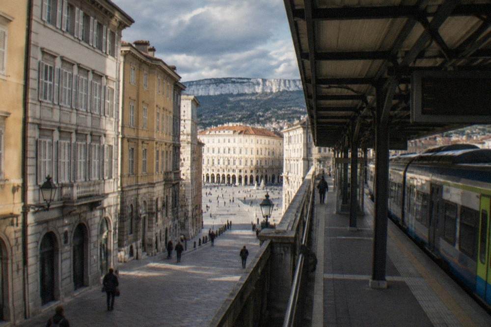 A regional train standing at Trieste Centrale station, Italy, with the platform canopy and departures board above it.