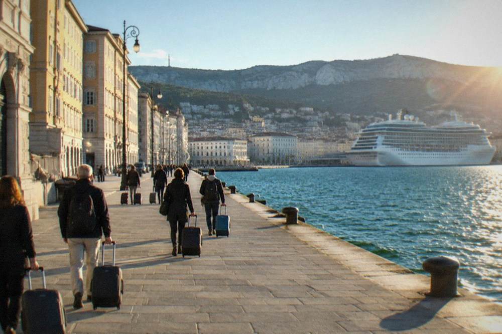 Passengers with suitcases walking along the Trieste seafront towards the cruise terminal, with the shoreline buildings behind them.