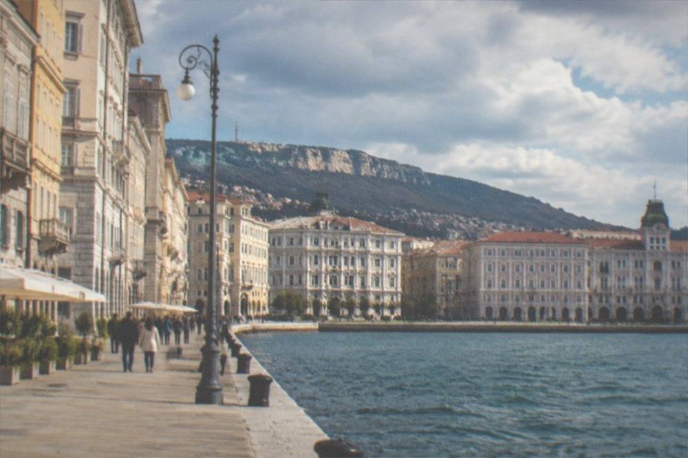 A wide seafront square in Trieste, Italy, with elegant historic facades and people walking beside the water.