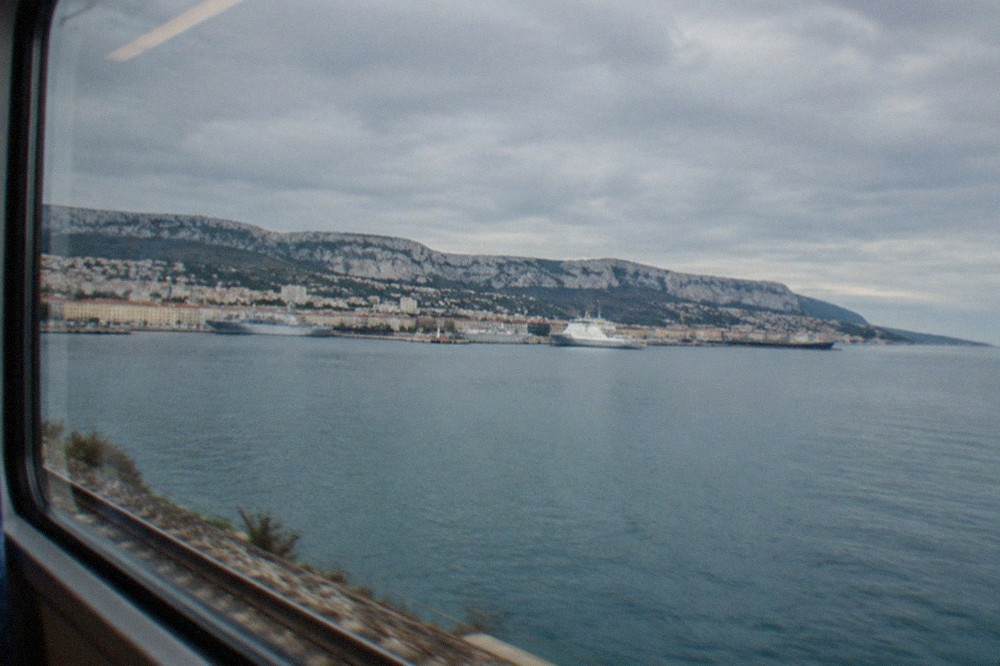 The Gulf of Trieste seen from a train window, with calm blue water, distant moored ships and pale limestone cliffs rising behind the shoreline