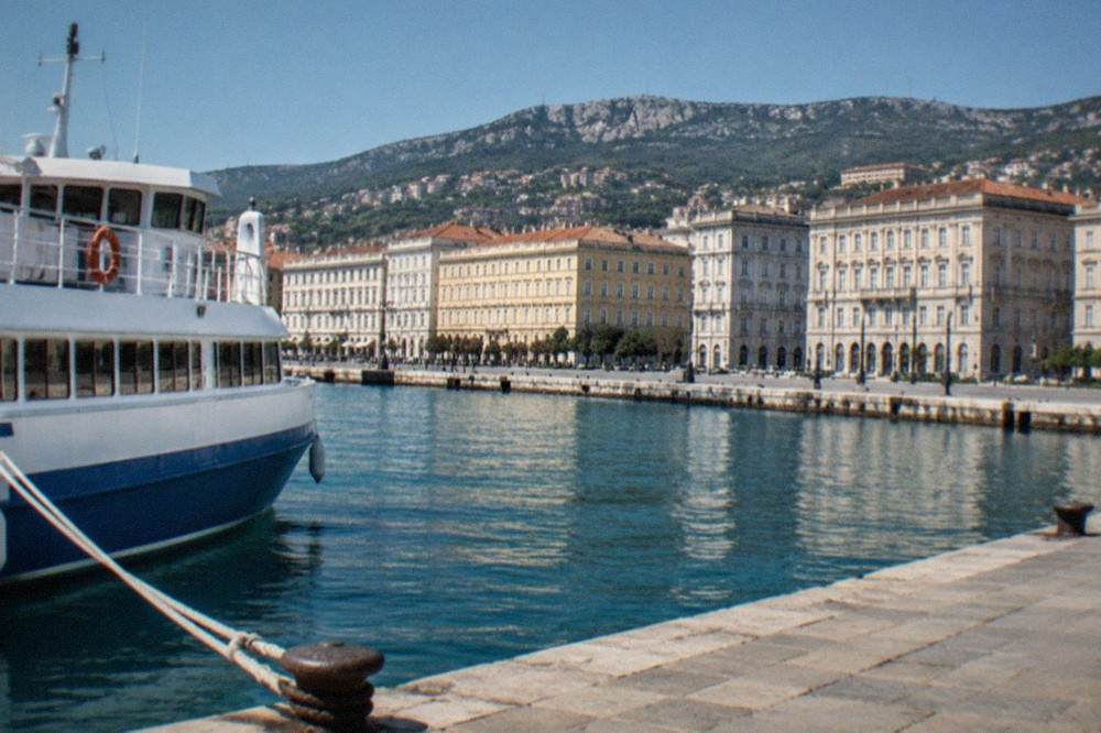 A passenger ferry tied up at a quay on the Trieste waterfront, with the historic seafront buildings in the background