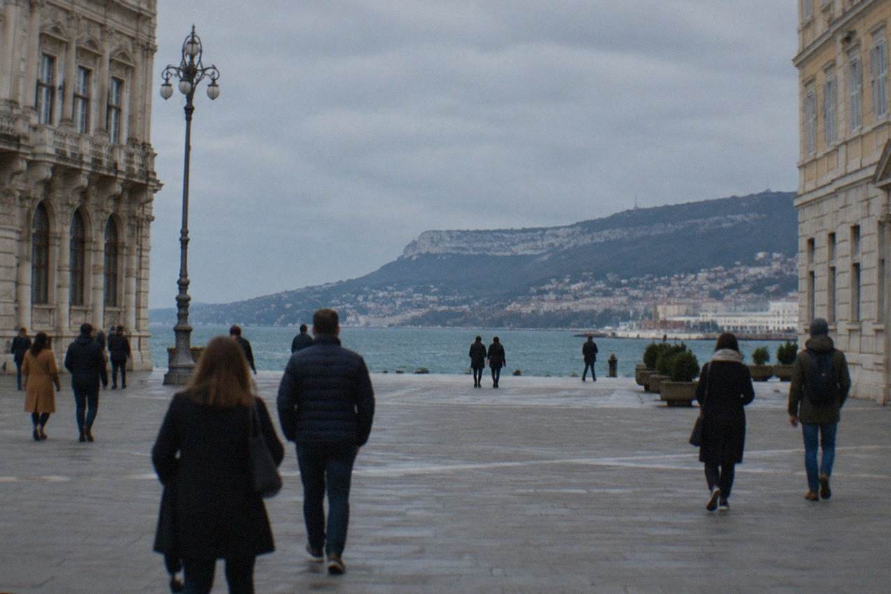 Pedestrians crossing a large seafront square in Trieste with ornate Habsburg-era facades and the Adriatic behind them