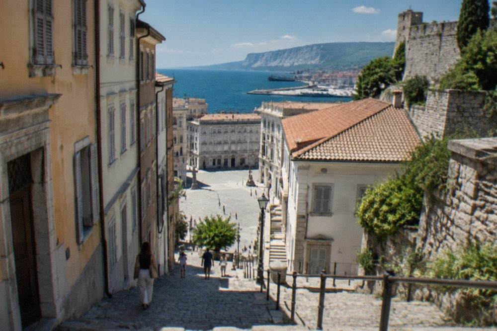 A stepped cobbled lane climbing a hillside in an old Italian port town toward a castle wall