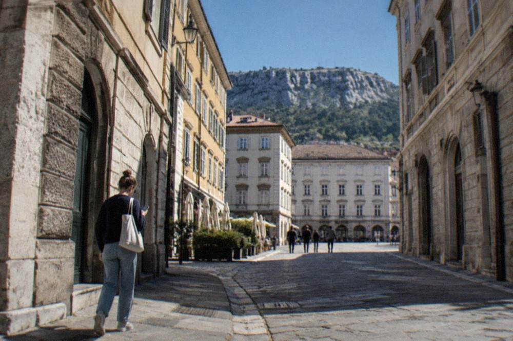 A self-guided walker reading a phone at a cobbled street corner in Trieste's old town