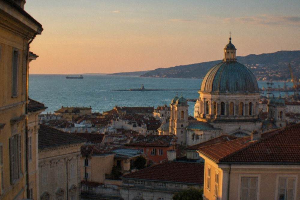 An evening view over Trieste's rooftops toward the Adriatic, with the dome of a historic church lit against the sky