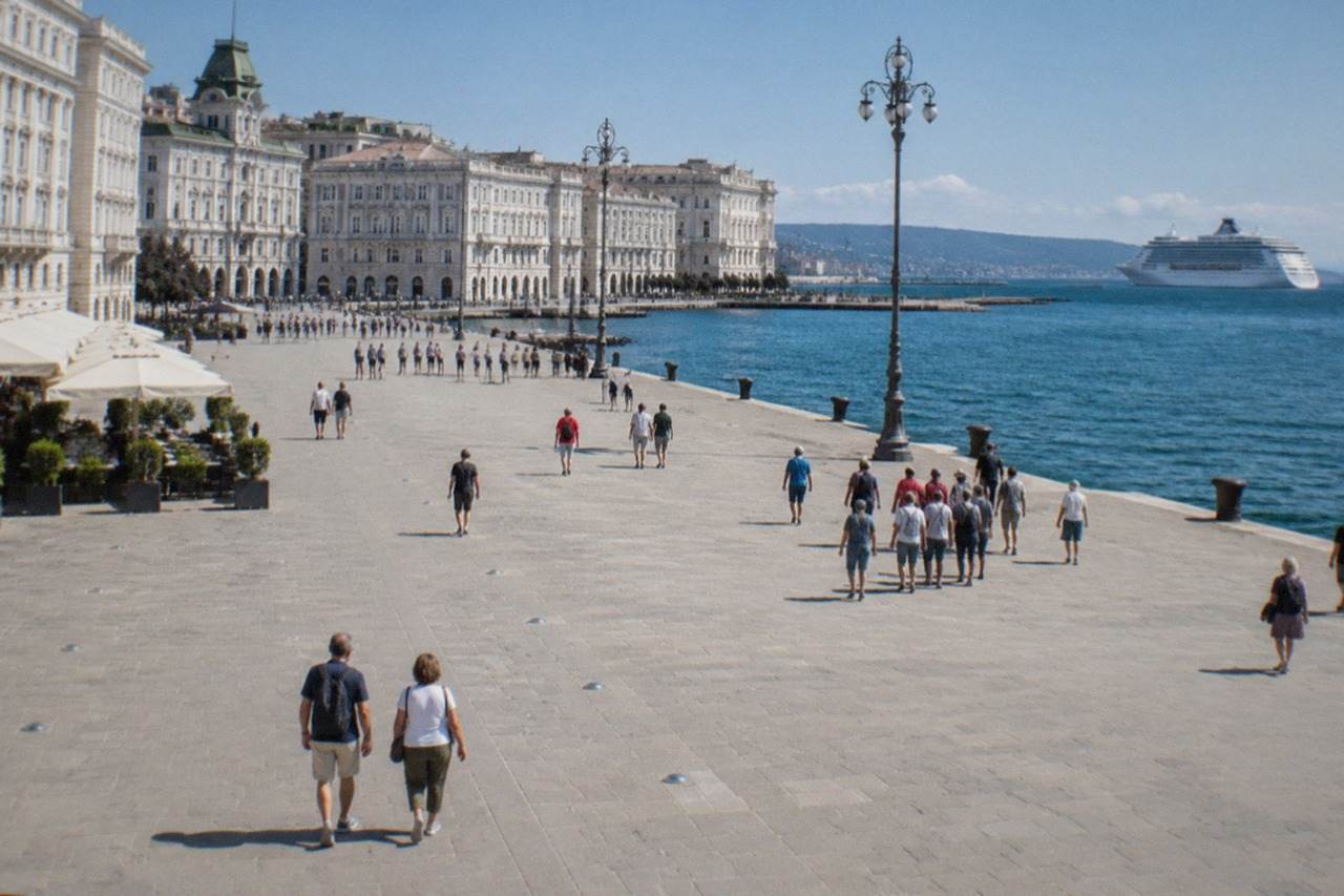 Cruise ship passengers walking along the Trieste seafront with the ornate waterfront buildings of Piazza Unità d'Italia behind them