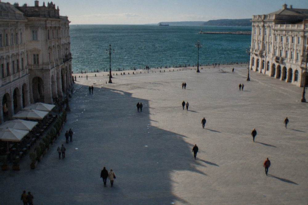 The wide open square of Piazza Unità d'Italia in Trieste with its ornate buildings facing the sea and a few pedestrians crossing the paving