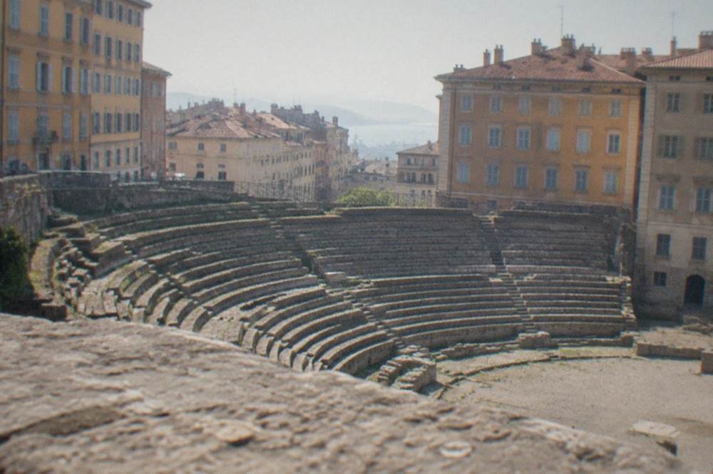 The Roman theatre of Trieste seen from the street, with old town buildings rising behind the ancient stone seating