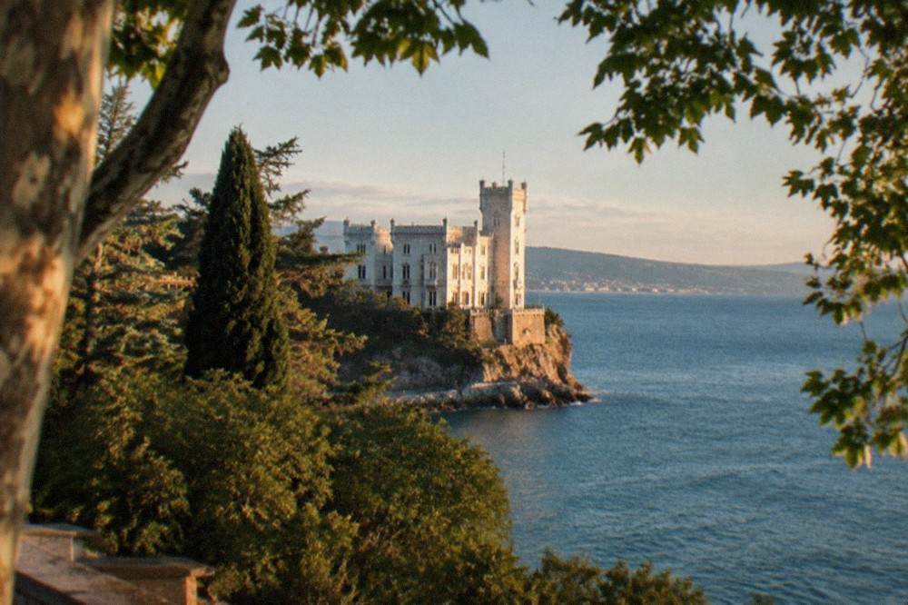 A white castle on a rocky headland above the Adriatic Sea near Trieste, Italy, seen through parkland trees