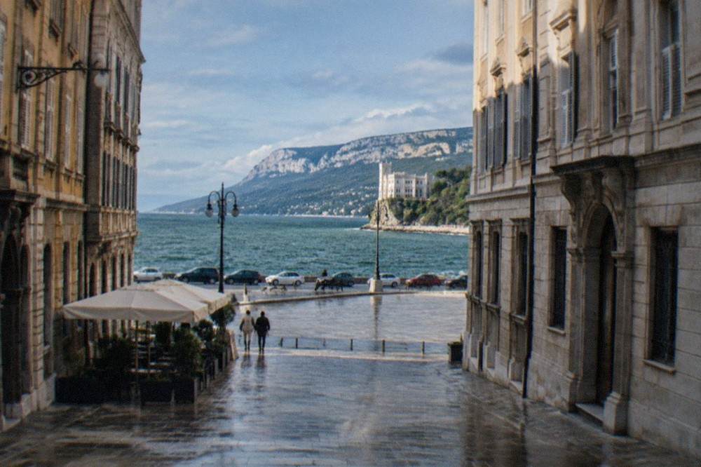 Cars on a coastal road above the sea with a white castle visible in the distance near Trieste