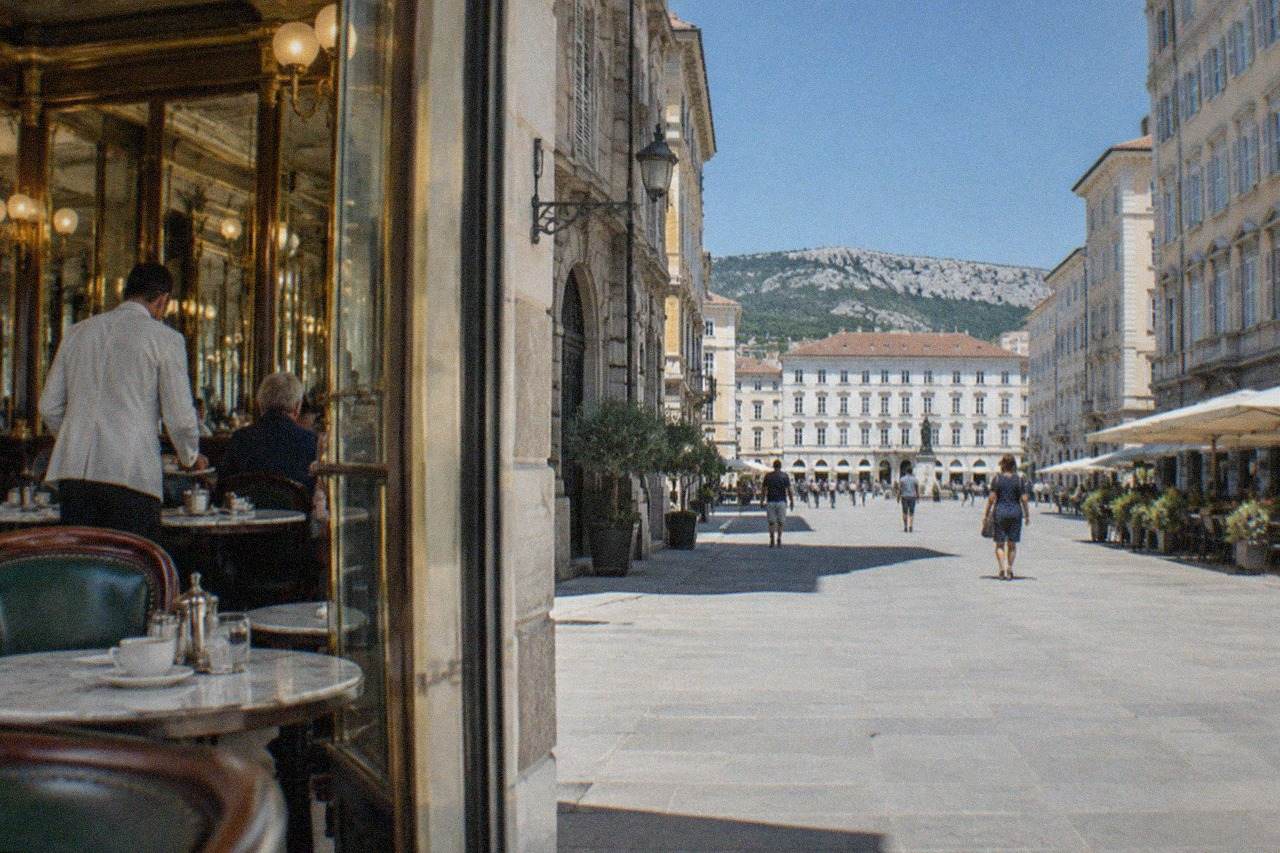 Waiters in white jackets serve coffee at marble tables inside an elegant historic Trieste cafe with brass fittings and mirrored walls