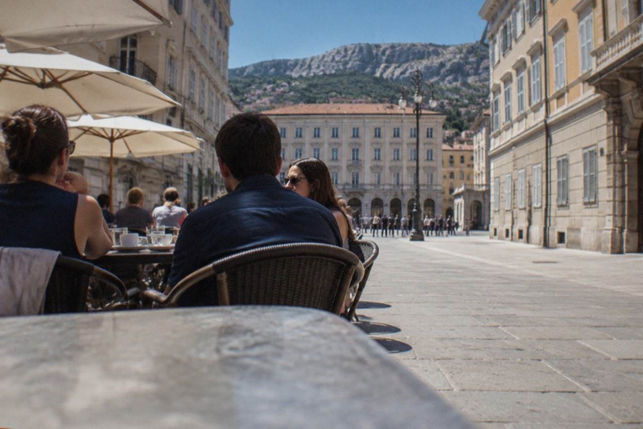 People talking at outdoor tables in a square in Trieste, Italy, with historic facades behind them