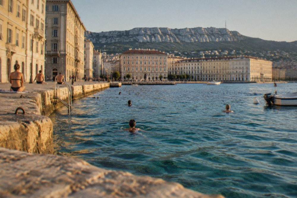Swimmers and small boats in clear water near a stone jetty on the Trieste coastline