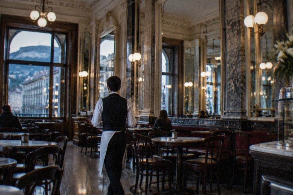 A waiter carrying a small tray of espresso cups through a grand mirrored café interior in Trieste