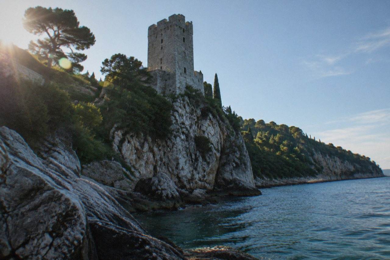 A stone castle tower standing above a rocky Adriatic shoreline with wooded cliffs behind it