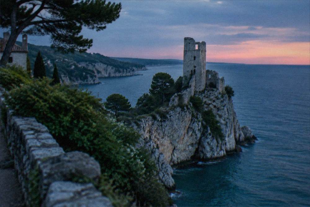 A ruined stone tower on a narrow rock spur above the sea at sunset