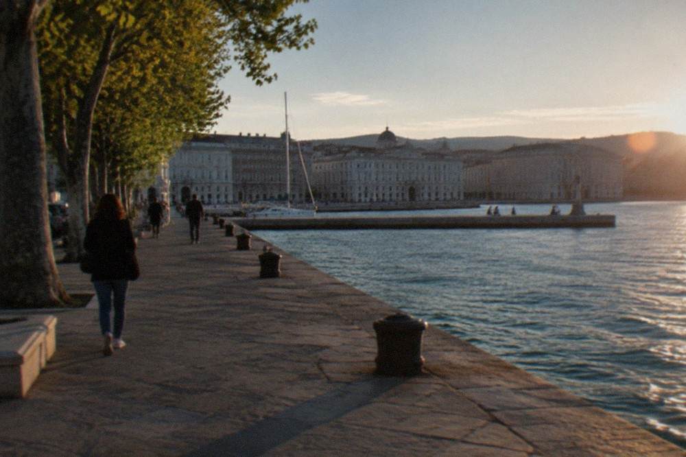 A stone pier reaching into a calm Adriatic gulf with a grand European square behind it