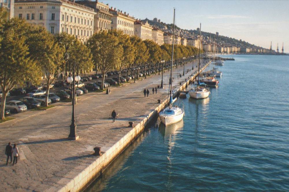 An evening view along a lit seafront promenade with the lights of a European port city reflected in still water