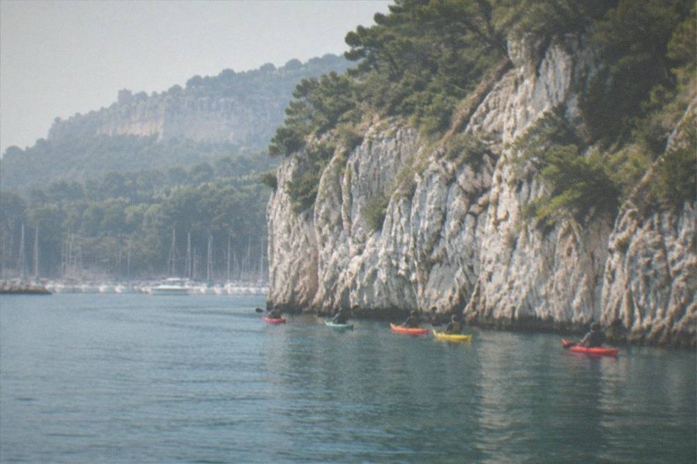 A line of sea kayaks paddling close beneath pale limestone cliffs above calm blue water