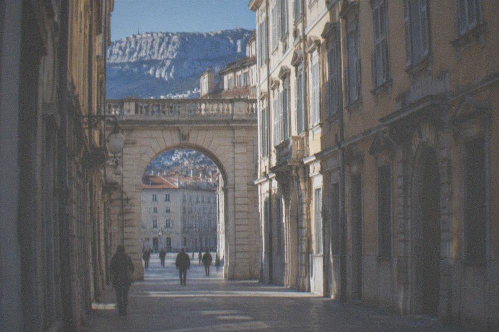 A sunlit stone archway with a blue sky above old Trieste rooftops