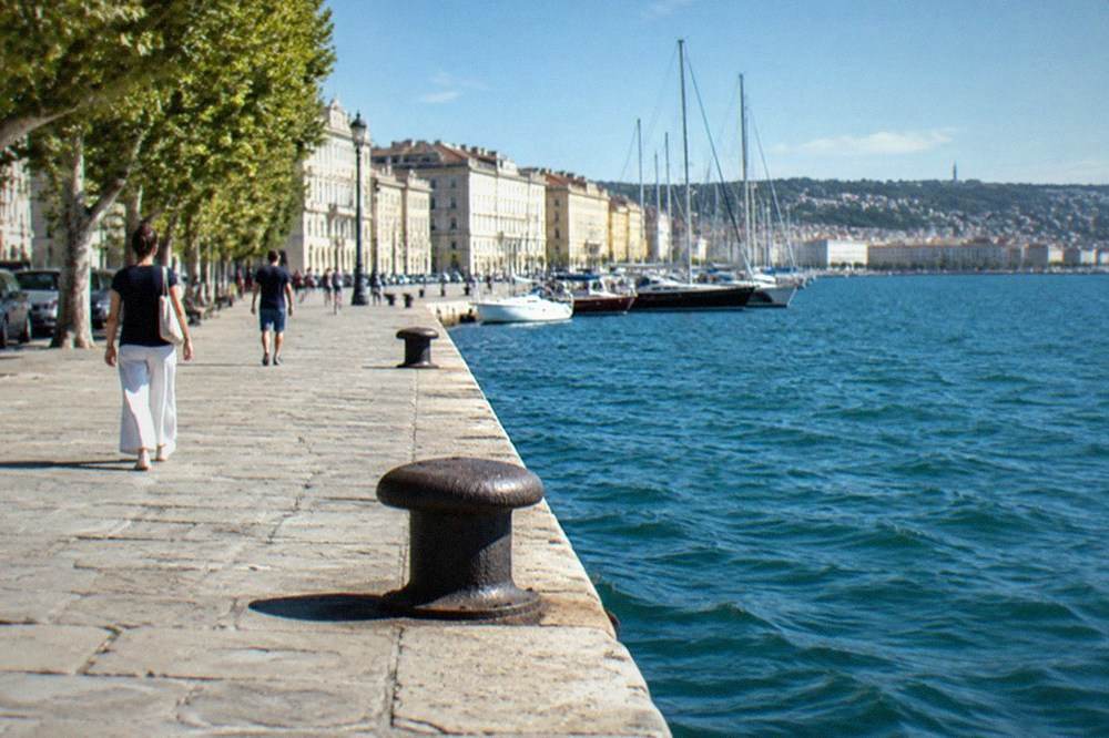 A paved seafront promenade in Trieste with people walking beside the Adriatic on a clear day