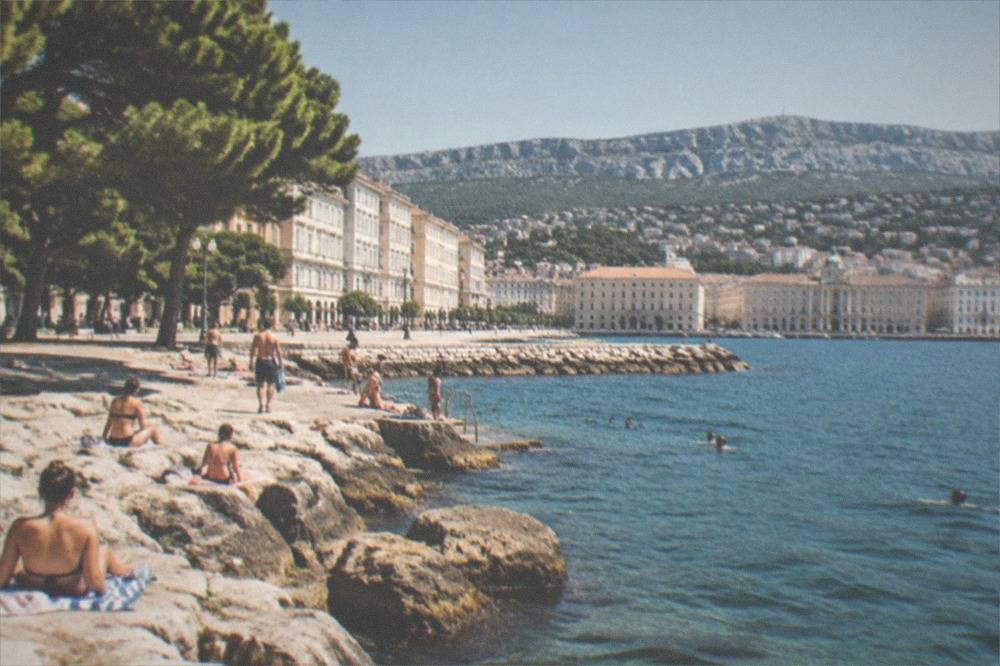 Bathers drying off on flat seaside rocks beside a stone bathing platform in Trieste