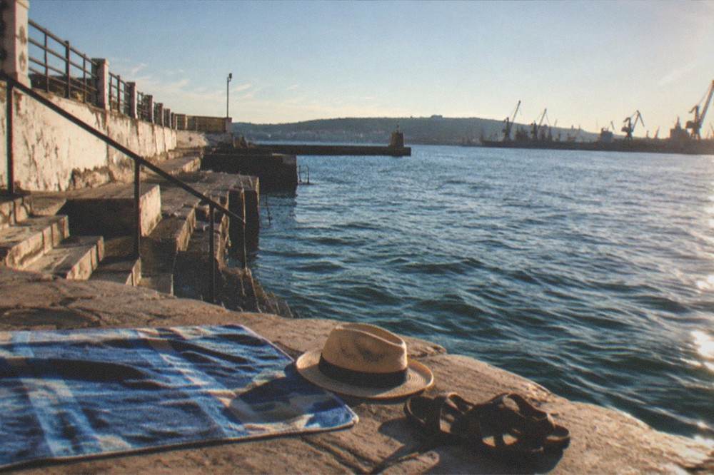 A swimmer's towel, hat and sandals laid out on a sunlit stone platform beside calm sea water