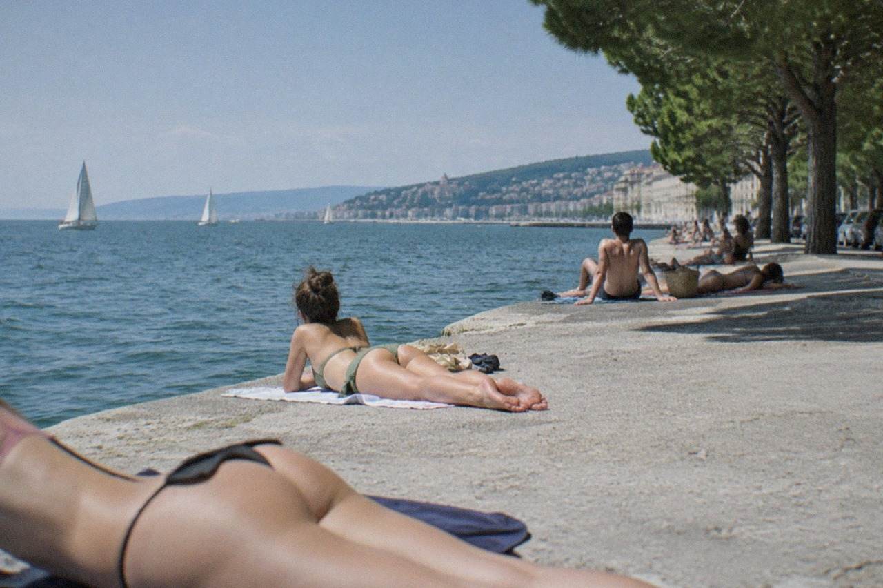 People sunbathing on the concrete seafront promenade at Barcola with sailboats out on the Gulf of Trieste