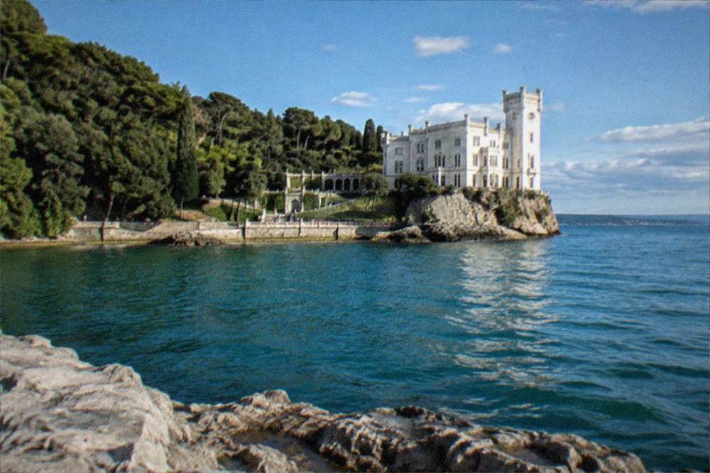 The wooded shoreline and white walls of Miramare Castle seen from the coastal path near Barcola