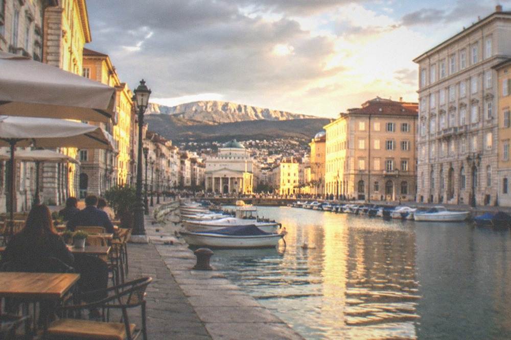 Outdoor café tables beside a city canal in Trieste at golden hour