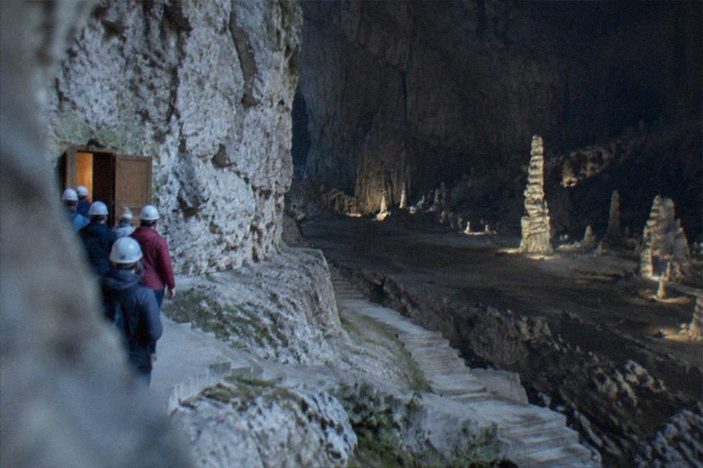 Visitors in helmets and walking shoes gathered at a cave entrance with a wooden door in a limestone cliff