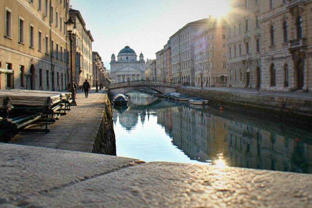 A narrow canal with ornate bridges and a domed church reflected in the water between tall buildings