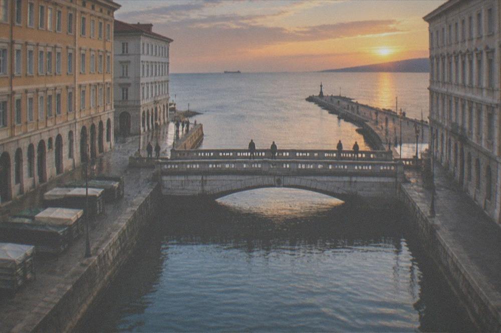 A long stone pier extending into a calm blue gulf with people walking at sunset