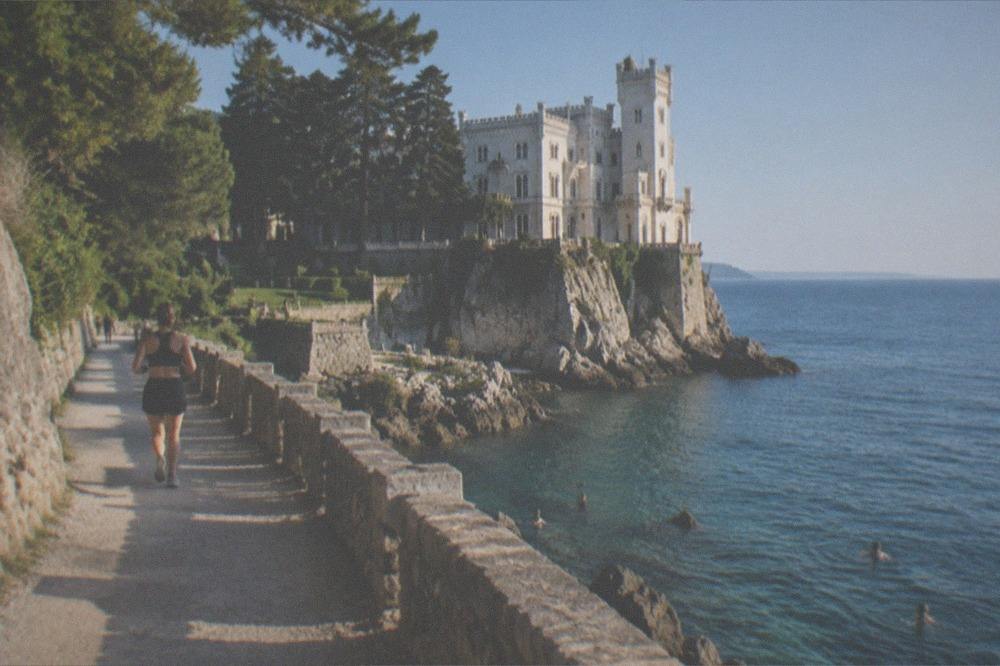 People swimming in clear water beside rocks below a castle headland