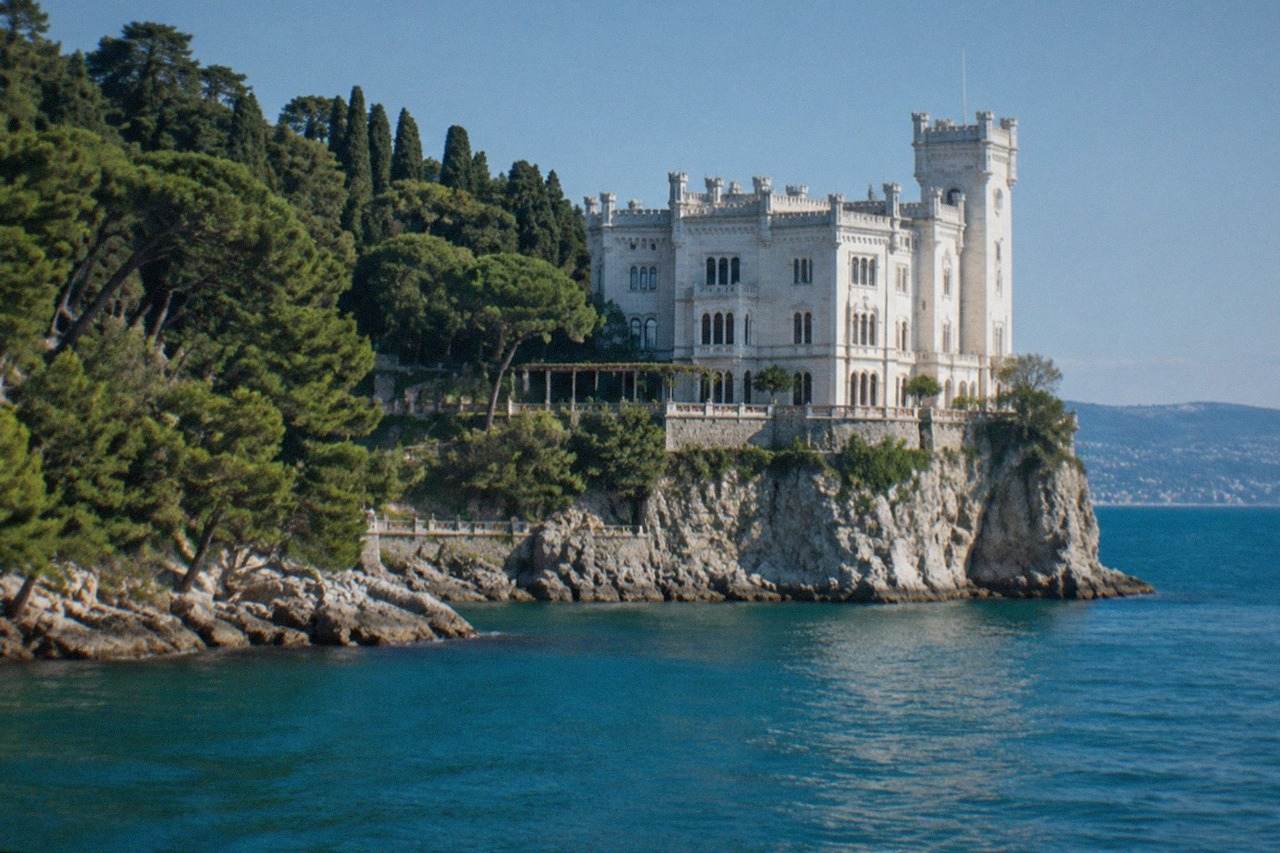 The white limestone facade of Miramare Castle rising above the sea on a rocky point with pine trees along the shore
