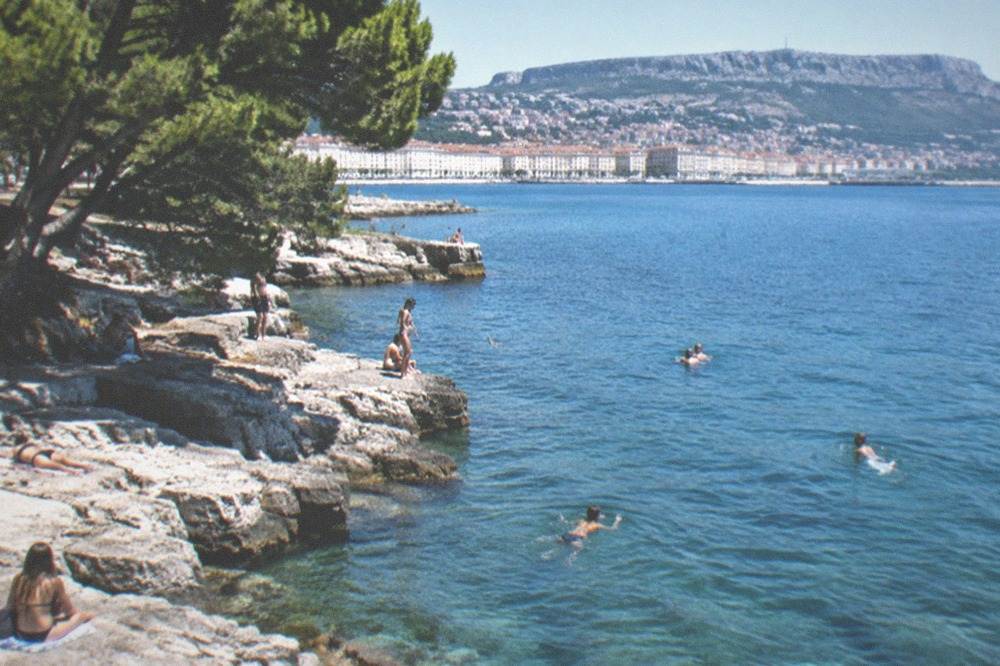 People swimming from flat limestone rocks into calm clear water on the Trieste coast in summer