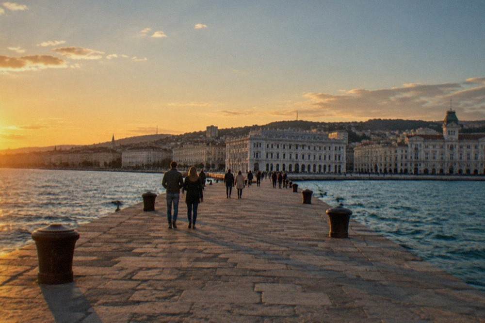 People strolling on a wide stone pier at sunset with a large square behind them