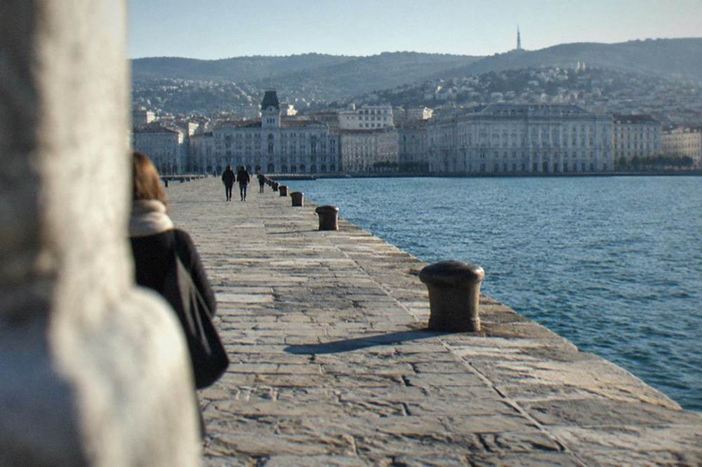 A wide seafront square meeting a calm sea, with a stone pier extending from the shore
