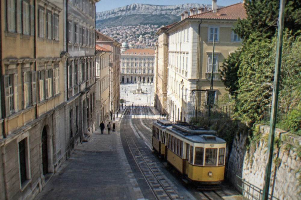 A historic two-car tram climbing through green hills above Trieste on a clear afternoon