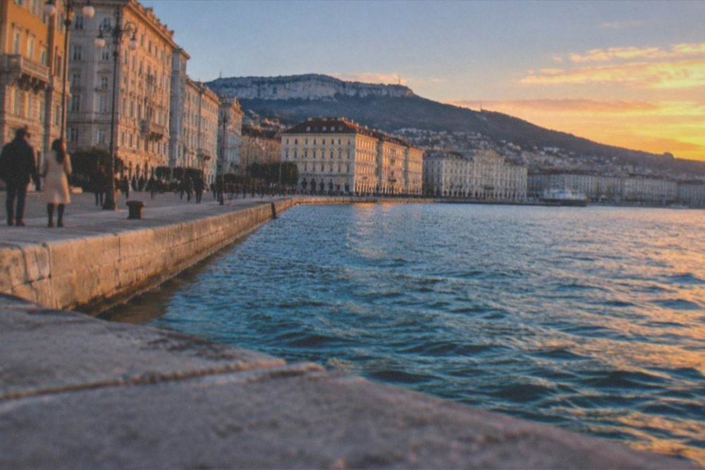 The Trieste waterfront at golden hour with the sea, historic facades and people strolling along the Rive