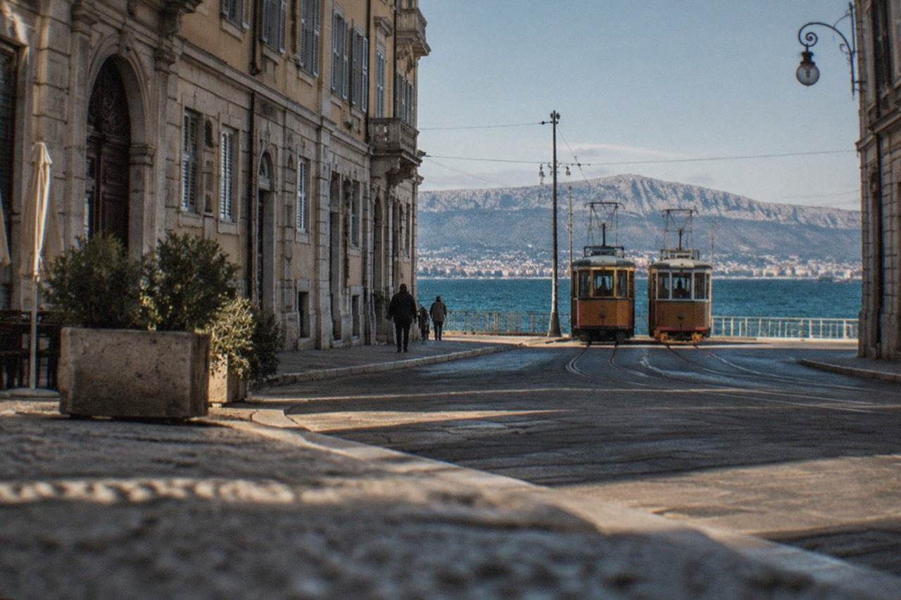 A vintage two-car tram standing at a street terminus in Trieste with the sea behind it