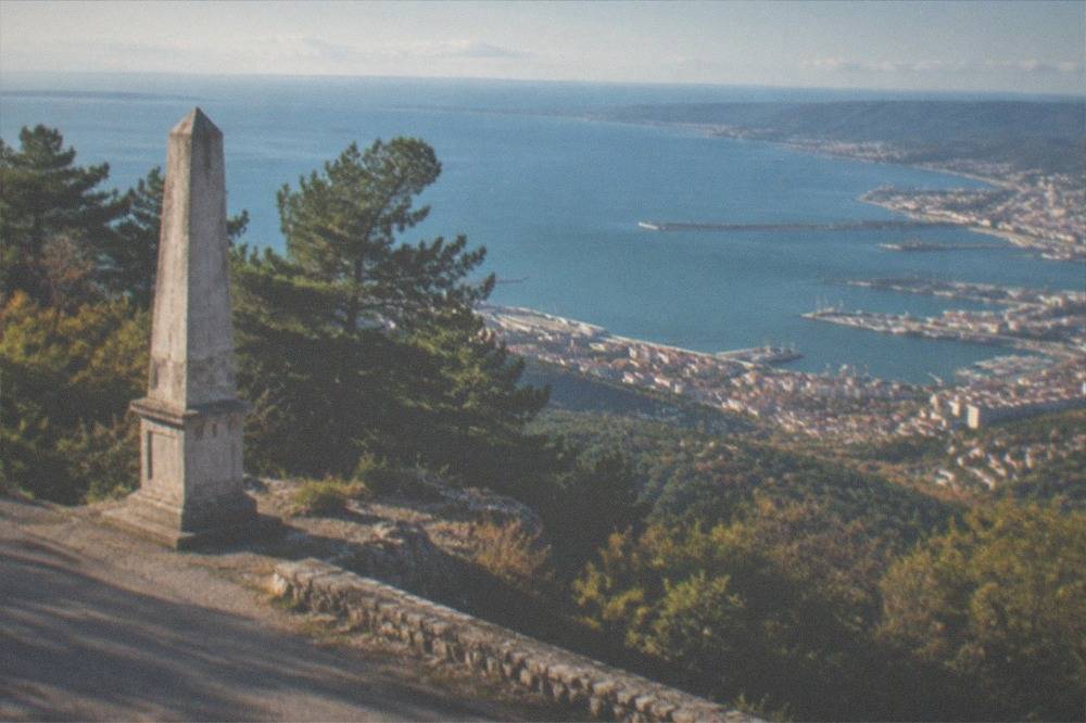 A stone obelisk beside a road at the edge of a plateau with a wide view over a bay and a city