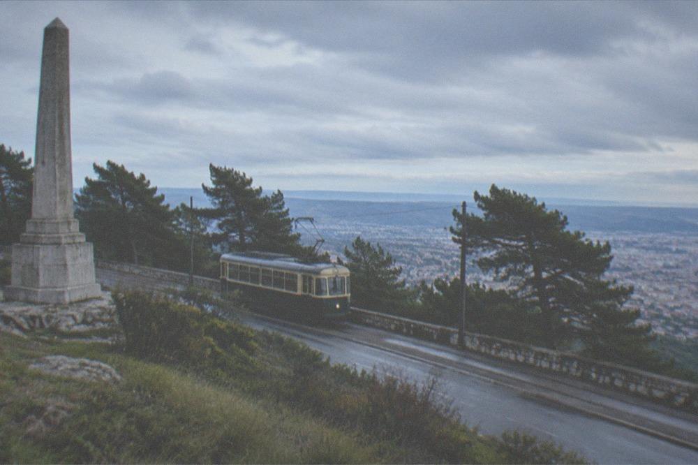 A tram on a high plateau road with wind-bent trees and a grey sky